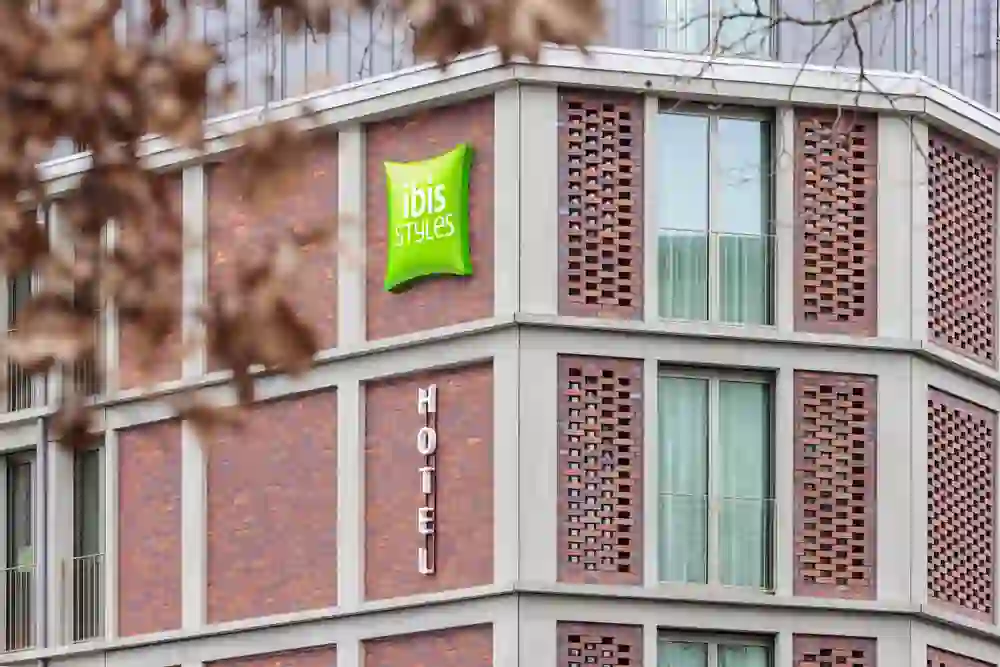 The picture shows the exterior façade of the Ibis Styles Hotel, characterised by a bright green billboard. The brick construction and large windows give the building an urban and modern character.
