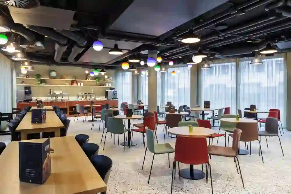 The photo shows the buffet of a modern bar or restaurant at the Hotel Ibis Styles Bern City with a relaxed and inviting atmosphere. Fresh fruit and pastries are on display, colourful chairs are grouped around wooden tables and the lighting creates a warm ambience.