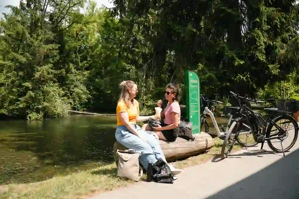 The picture shows two people chatting on a tree trunk on the bank of a river or lake. The person on the left is wearing a yellow top and jeans, while the person on the right is wearing a pink T-shirt and dark trousers. Behind them are several e-bikes attached to a sign with a map and information. The background is characterised by lush green trees and the clear water of the river or lake. It appears to be a sunny day and the scene looks relaxed and peaceful.