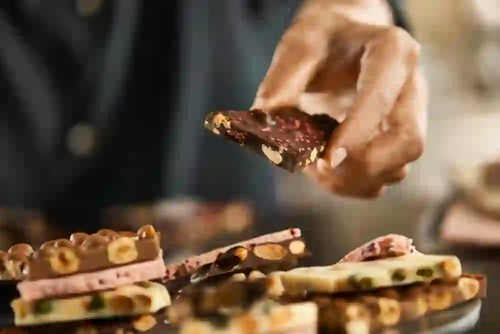 Eine Hand hält ein verlockendes Stück Schokolade von Läderach, umgeben von einer Auswahl an feinsten Schokoladenkreationen, die Genuss und Handwerkskunst suggerieren.