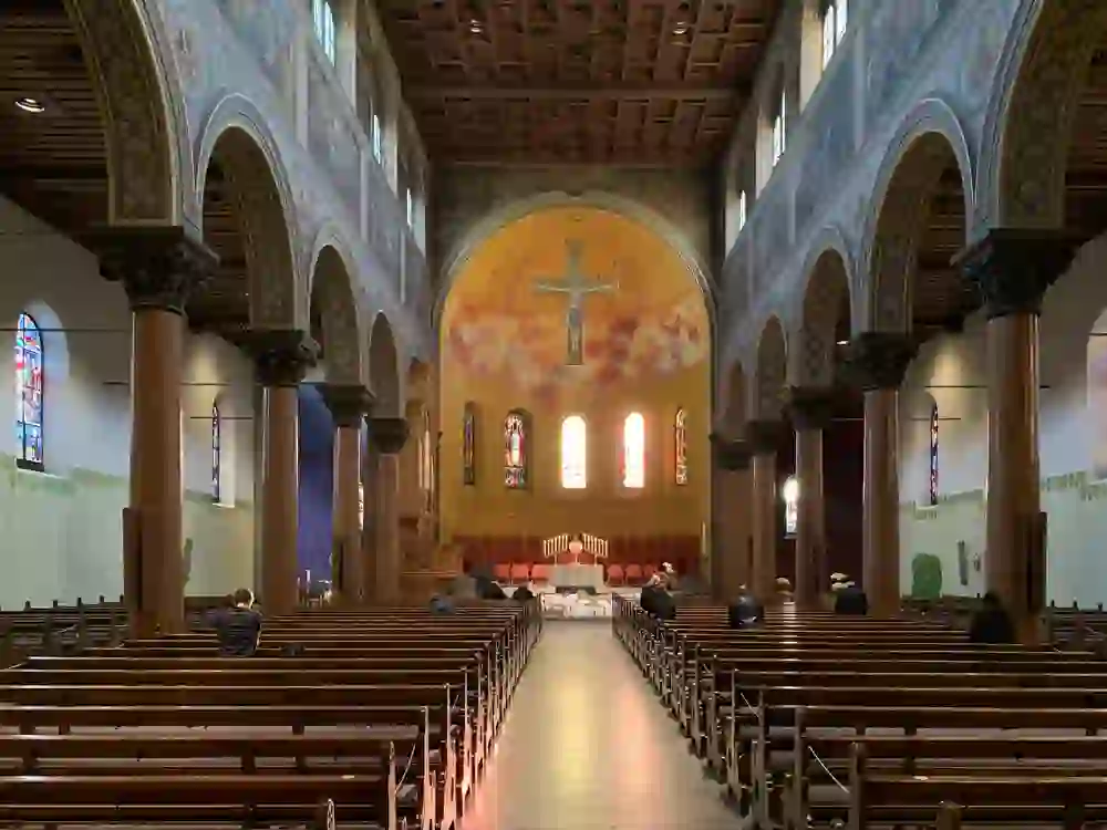 Das Bild zeigt das Innere der Dreifaltigkeitskirche Bern mit einer traditionellen Kirchenarchitektur. Die Decke ist mit Holzbalken versehen, und es gibt eine Reihe von Säulen auf beiden Seiten des Mittelgangs. Bunte Glasfenster lassen natürliches Licht herein. Der Altar am Ende der Kirche ist gut beleuchtet, und es gibt einige Personen, die in den Bänken sitzen oder durch die Kirche gehen, möglicherweise in stiller Andacht oder Besichtigung.