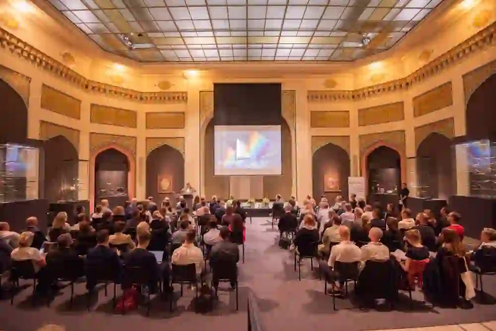 Der orientalische Saal des Bernischen Historischen Museums fängt eine Atmosphäre konzentrierter Aufmerksamkeit ein, während das Publikum einer Präsentation folgt, umgeben von prächtigen dekorativen Elementen, die eine kulturelle Brücke schlagen.