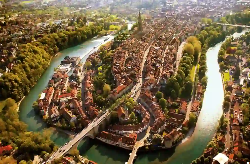 The image shows a drone shot of Bern with the River Aare meandering through the historic old town. The warm sun bathes the rooftops in a golden light and creates a peaceful, picturesque atmosphere