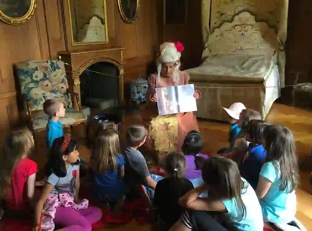 In a historic-looking room, children listen intently to a storyteller. They sit on the floor, surrounded by antiques that seem to reflect the times. A feeling of curiosity and childlike wonder fills the air.