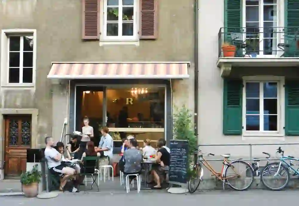 The image captures a lively outdoor café scene where people gather in a relaxed atmosphere in sunny weather. The warm atmosphere is enhanced by the historic architecture and passing bicycles, creating a sense of community and serenity.