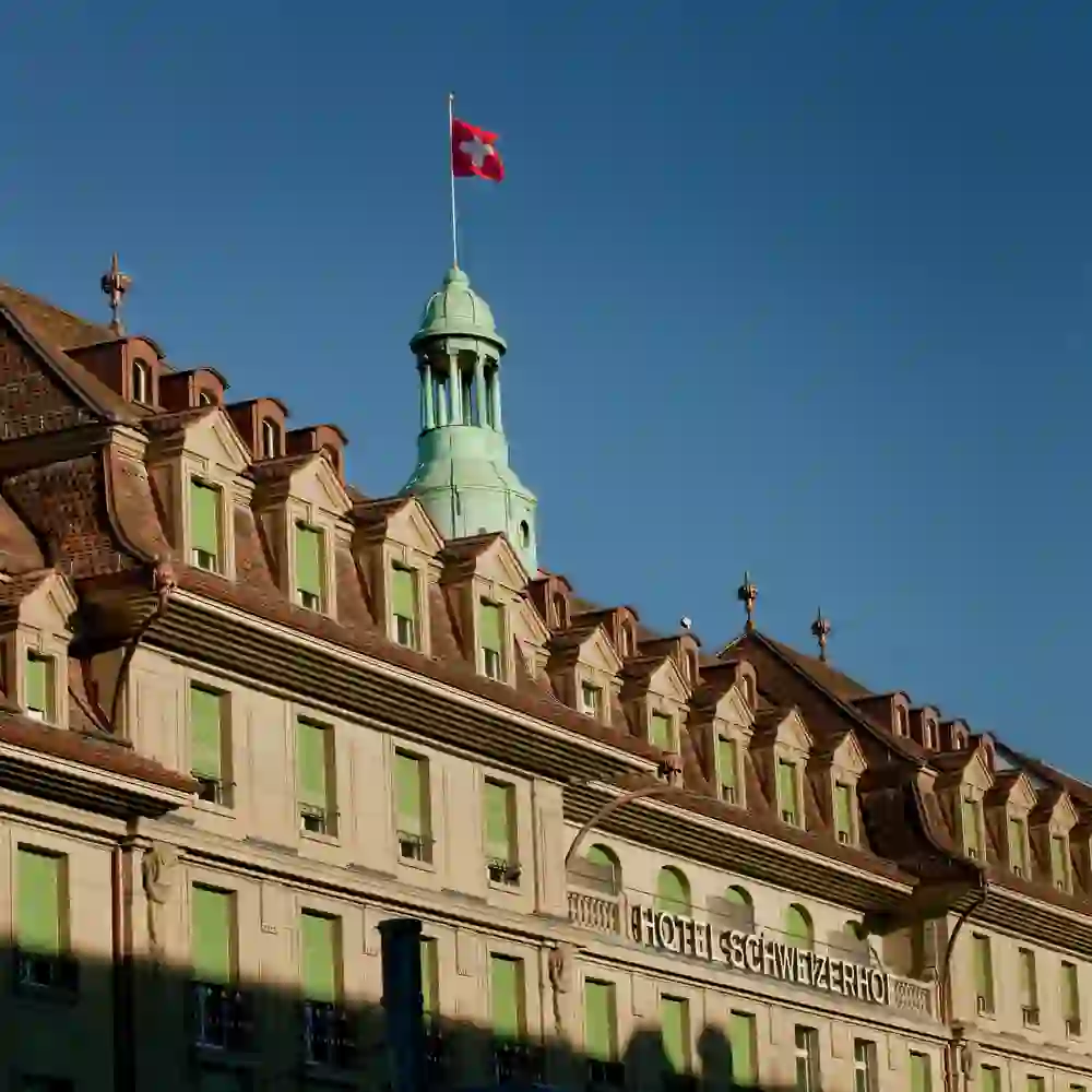 Das Bild zeigt das Hotel Schweizerhof in Bern in warmem Sonnenlicht, das eine ruhige und einladende Atmosphäre erzeugt. Die stolze Schweizer Flagge weht hoch über dem klassisch eleganten Gebäude, ein Symbol für Gastfreundschaft und Tradition.