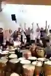 Eine fröhliche Gruppe lächelt in die Kamera, umgeben von Trommeln bei einem Teamevent mit Musik. Ihre begeisterten Gesichter und erhobenen Hände spiegeln Gemeinschaftsgeist und Freude am gemeinsamen Rhythmus wider.