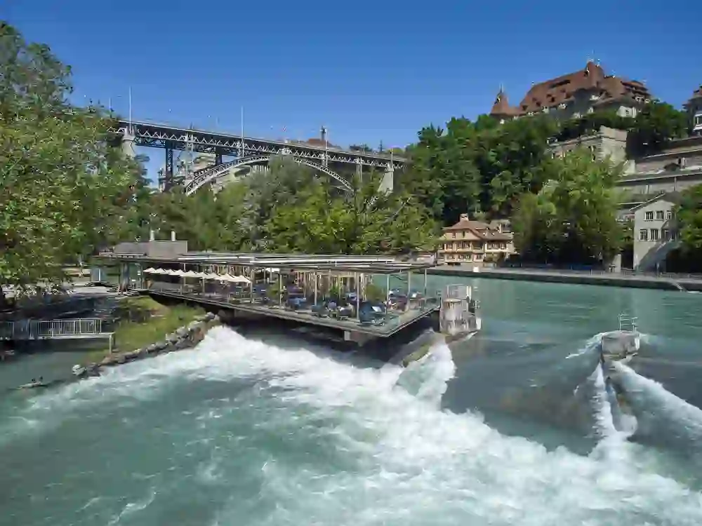 The picture shows a restaurant on the River Aare, below a bridge and next to a waterfall. You can see a terrace directly on the water. The atmosphere is dynamic and refreshing, characterised by the flowing, turquoise-coloured water. A castle or historic building rises up in the background, surrounded by trees. It seems to be a sunny, clear day, perfect for guests who want to enjoy nature and the view.