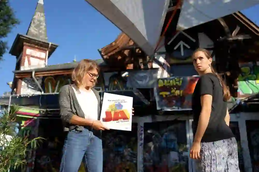 Two women stand in front of the Reitschule in Bern, which is characterised by urban street art. One of them is holding a poster with a message, while the other looks seriously into the distance. They are on a city tour about women's suffrage, where they want to draw attention to something important.