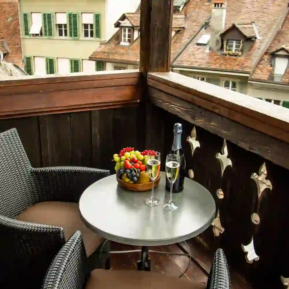 Das Bild zeigt eine charmante Balkonszene mit Blick auf traditionelle Dachfenster der Berner Altstadt. Auf dem Tisch steht eine Schale mit frischem Obst neben zwei Gläsern und einer Flasche Sekt, was eine festliche und gemütliche Atmosphäre schafft. Die Sitzgelegenheiten laden zum Verweilen und Genießen der Aussicht ein.