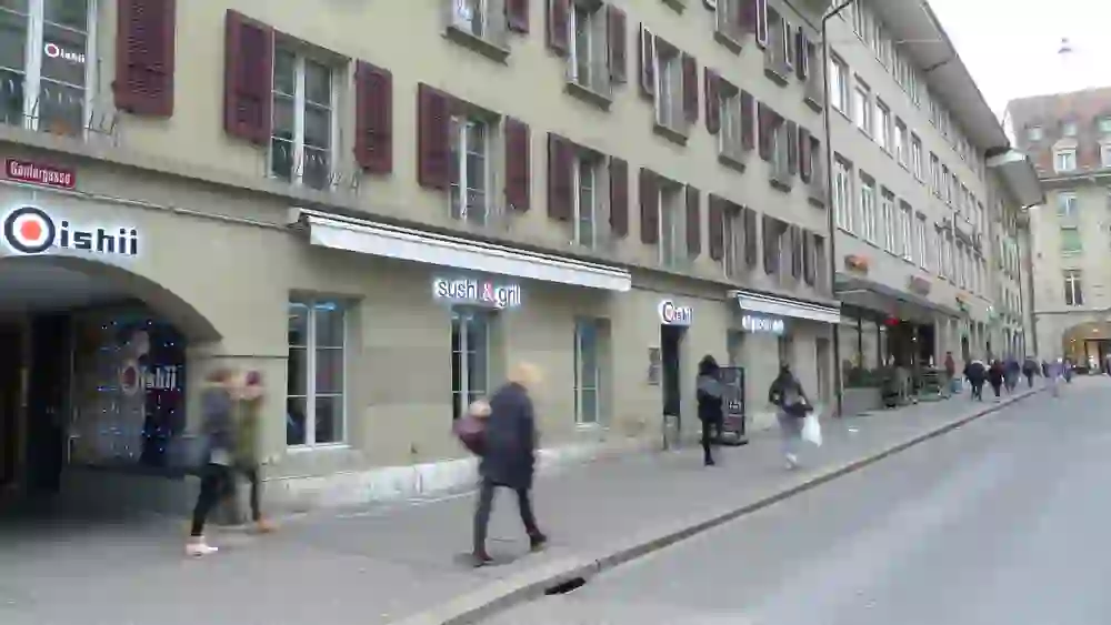 L'image montre l'extérieur du restaurant Oishii Sushi & Grill à Berne. La scène semble animée et dynamique, avec des gens qui passent à la hâte malgré le temps maussade, ce qui indique une ville animée. Les couleurs sont atténuées, mais l'enseigne lumineuse du restaurant offre un point de mire lumineux.