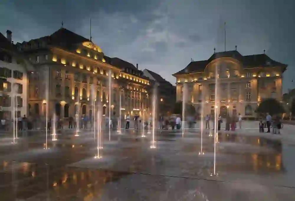 L'immagine mostra la Bundesplatz di Berna al tramonto con fontane, edifici illuminati e persone in piazza.