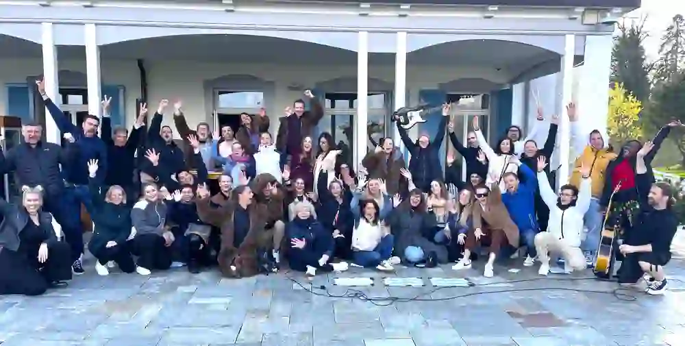 Un gruppo esuberante posa ridendo per una foto durante un evento musicale di gruppo. Volti gioiosi e posture energiche sono visibili ovunque, e alcuni hanno in mano degli strumenti. La scena è piena di entusiasmo e di spirito di squadra.