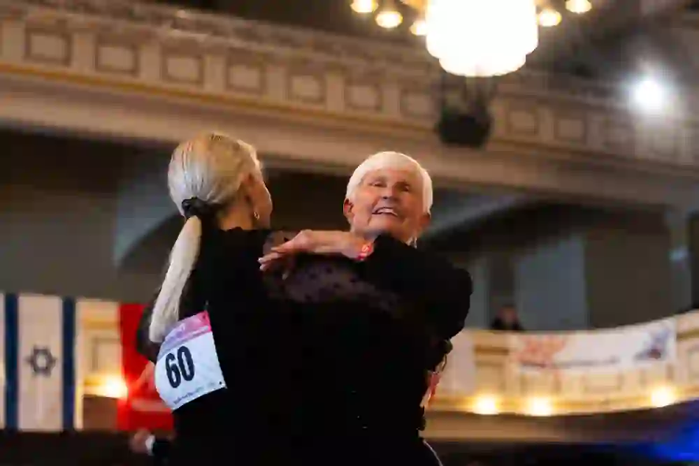 Das Bild zeigt zwei Menschen in festlicher Kleidung, die miteinander tanzen. Eine ältere Dame mit einem breiten Lächeln und einem schwarzen Kleid strahlt Freude aus, während sie mit ihrer Partnerin mit langem, blondem Haar tanzt. Die Atmosphäre wirkt ausgelassen und freudig.