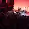 The picture shows an impressive live show in front of a packed audience. The stage is bathed in warm red light, on which a band is playing while the audience listens attentively. The two-storey hall offers the audience the opportunity to enjoy the concert from different perspectives. The intense lighting and dense atmosphere create a sense of community and enthusiasm for the music, while the silence in the room suggests a concentrated, reverent mood.