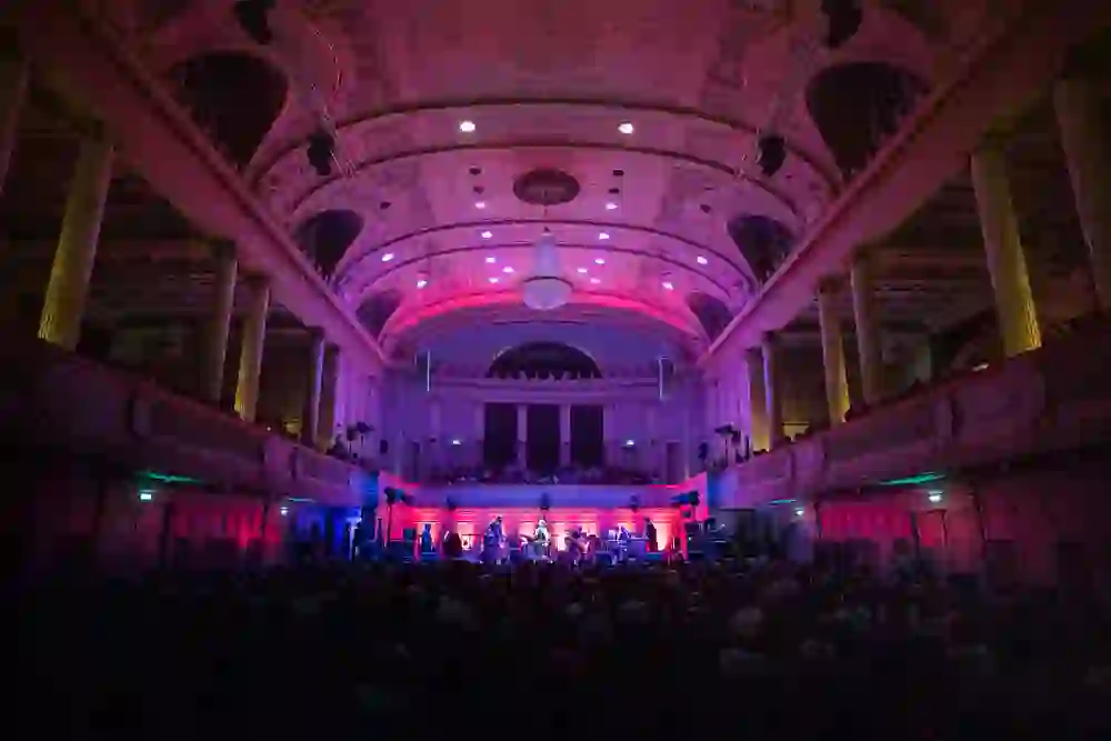 L'immagine mostra una scena di concerto nella Sala Grande del Casinò di Berna, dove il pubblico è immerso nella luce violetta del palco e del lampadario luminoso. L'architettura della sala crea un'atmosfera imponente ed elegante che enfatizza l'esecuzione musicale.