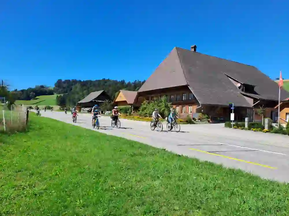 L'immagine mostra una scena rurale nella valle dell'Emmental, con diverse persone in bicicletta su una strada. La strada passa accanto alle tradizionali cascine svizzere, ben tenute e circondate da fiori e giardini. Sullo sfondo si intravedono colline e foreste. Il cielo è limpido e azzurro, a indicare una giornata di sole. Sulla sinistra dell'immagine c'è un prato verde con una recinzione. La scena appare tranquilla e idilliaca e le persone sembrano godersi la loro passeggiata in bicicletta.