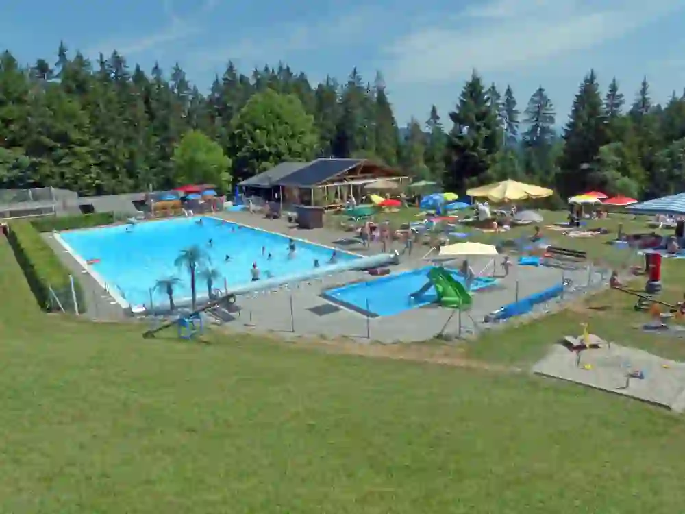 The picture shows the Eywald outdoor pool in the Gantrisch Nature Park. In the foreground is the 25-metre swimming pool, where several people are swimming. To the right of the large pool is a smaller children's pool with a slide. Around the pools, many people are lying on towels and under parasols to enjoy the sun. In the background you can see a small building that houses the outdoor pool's restaurant. The entire area is surrounded by a well-tended lawn and a wooded area. The atmosphere is relaxed and family-friendly.