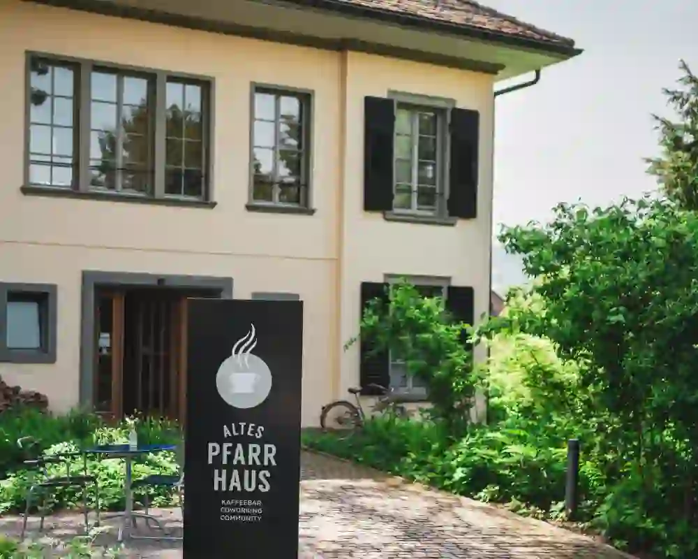 Vor einem historischen Haus steht das Schild einer Kaffeebar, umgeben von viel Grün und einem ruhigen Gartenweg. Die Szene wirkt einladend und entschleunigt, sie vermittelt Geborgenheit, Kreativität und einen Ort zum Ankommen und Verweilen.
