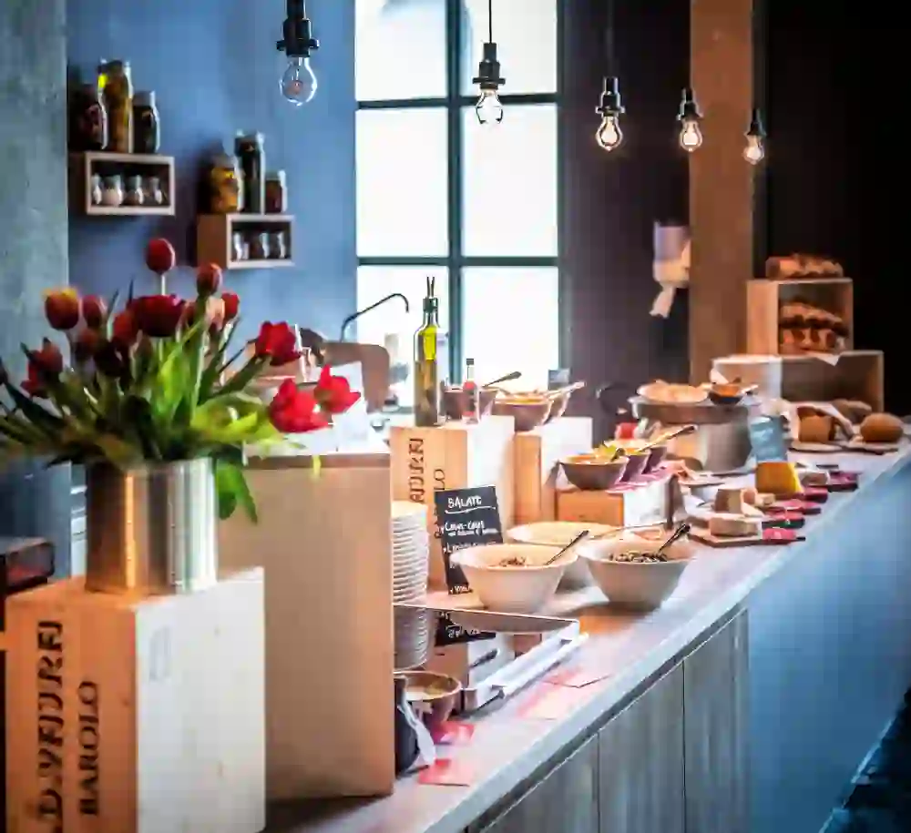 The picture shows a brunch buffet in a stylishly furnished room. Fresh flowers in a vase, various cereal bowls, cheese, bread and pickled food adorn the counter. The pendant lights and the large window create a warm and inviting atmosphere.