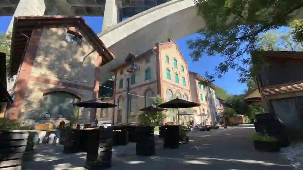 L'image montre l'extérieur de Chillfood's Füüri, un lieu accueillant qui peut être réservé pour des événements. Sous un impressionnant pont en béton se trouvent des bâtiments historiques en briques, complétés par des espaces modernes pour s'asseoir et des parasols, ce qui crée une atmosphère unique alliant charme industriel et hospitalité chaleureuse.