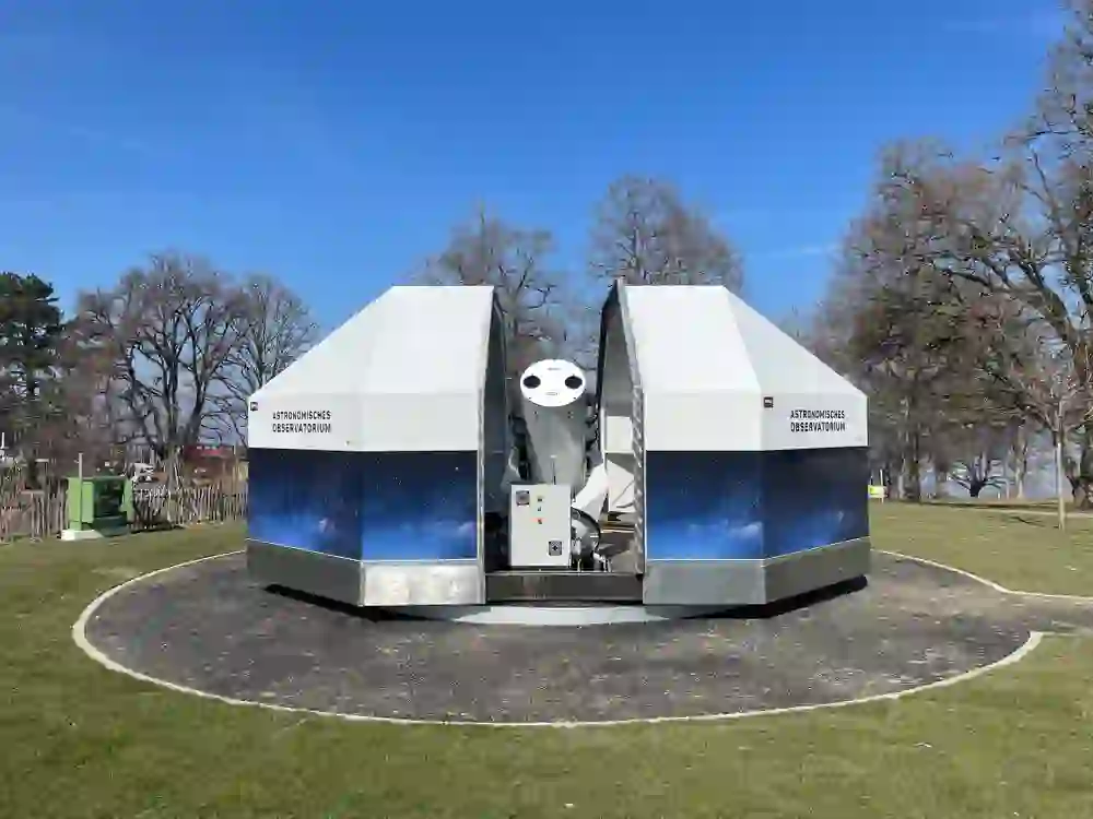 Ein modernes Observatorium steht bereit unter einem blauen Himmel, ein Tor zur Erkundung der Sterne, eingebettet in den ruhigen, grünen Gurtenpark.
