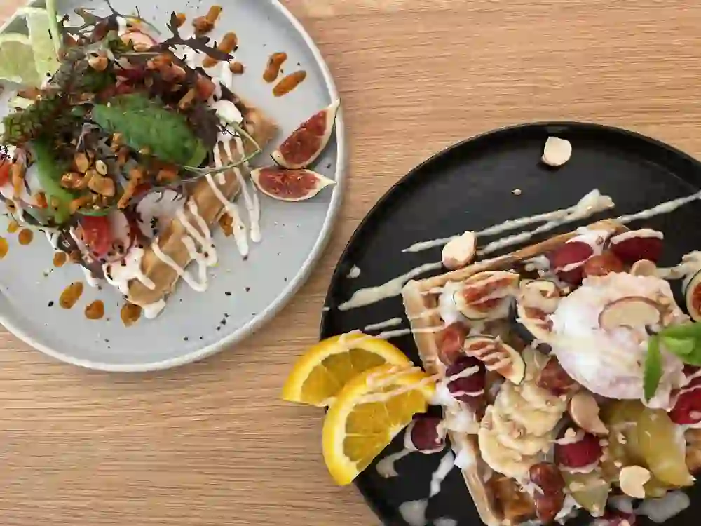 Das Bild zeigt zwei kunstvoll angerichtete Desserts auf Holztischen, die eine warme und einladende Atmosphäre schaffen. Links Waffeln mit Feigen und Creme, rechts Waffeln mit Früchten und Nüssen, beide ansprechend dekoriert, vermitteln Freude und Genuss.