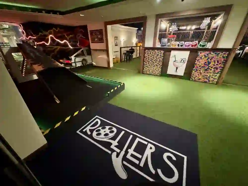 Innenansicht von Rollers Bern mit einer kreativ gestalteten Minigolfbahn im „Zurück in die Zukunft“-Stil. Links ein Autoteil mit Neonschrift, rechts die Bar mit bunten Golfbällen und farbigen Sonnenschirmen. Auf dem Boden ein Teppich mit dem Rollers-Logo.