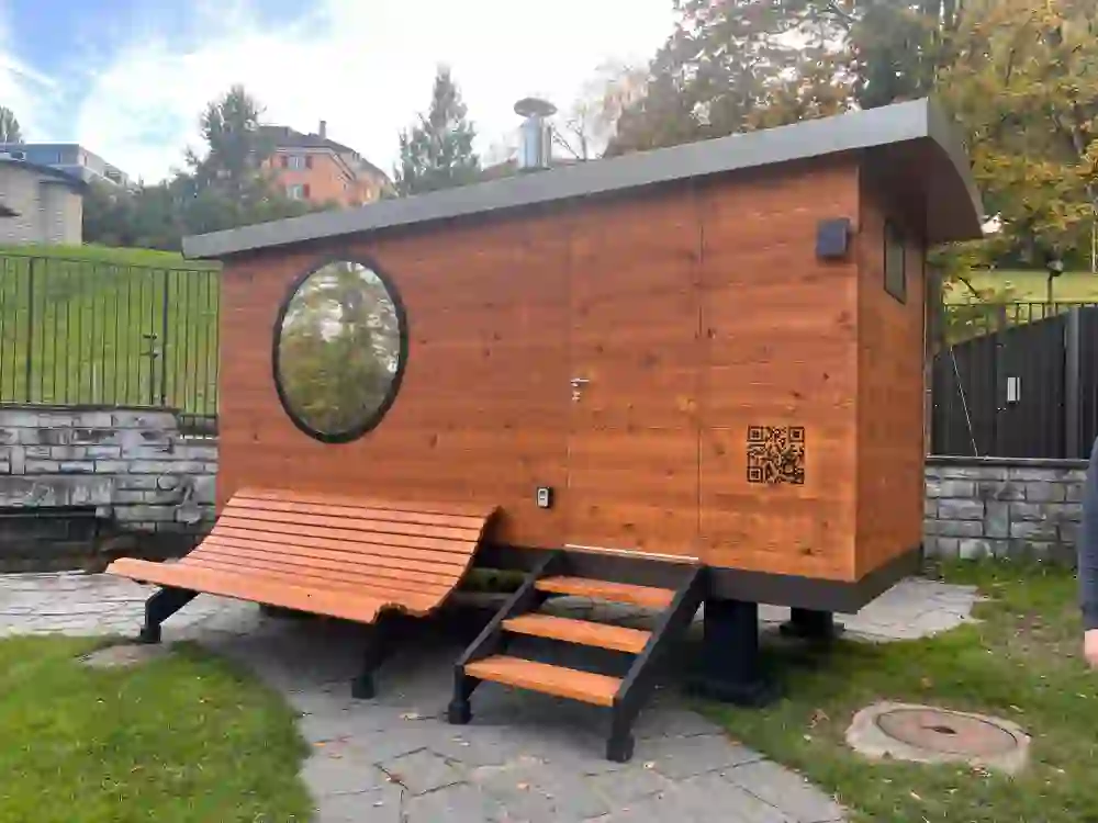 L'immagine mostra una moderna sauna all'aperto in legno caldo. L'apertura ovale della finestra e le sedie a sdraio in legno curvato davanti ad essa conferiscono un'aura invitante e rilassante. Circondata dalla natura, la scena invita a rallentare e a sentirsi bene.