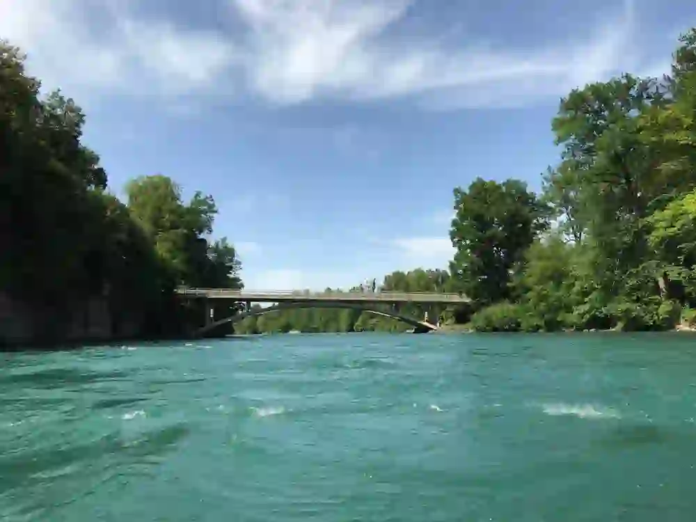 Das Foto präsentiert die Jabergbrücke in Kiesen, die sich malerisch über die Aare spannt. Umgeben von üppigem Grün, reflektiert das klare, türkisfarbene Wasser den blauen Himmel. Die Szenerie ist ruhig und idyllisch, ein perfekter Ort für Naturliebhaber und eine Oase der Ruhe. Die Brücke verbindet nicht nur zwei Ufer, sondern symbolisiert auch die Verbindung von Technik und Natur in harmonischer Koexistenz.