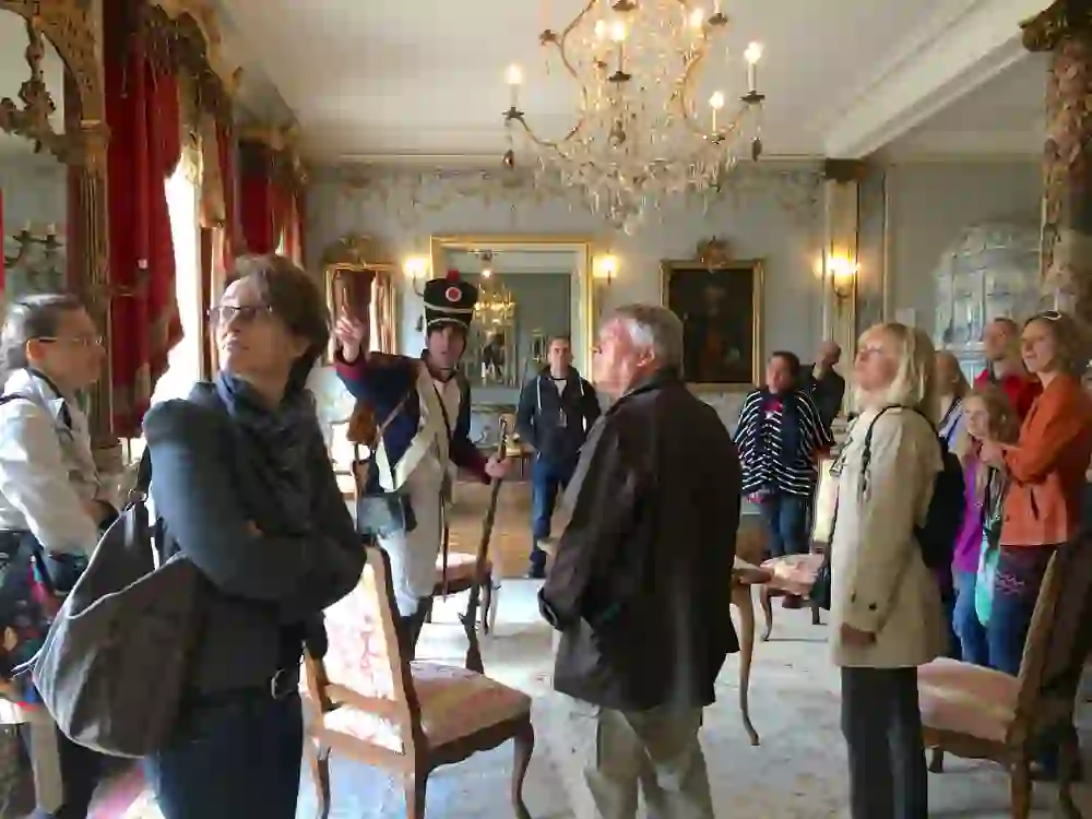 In einem prachtvollen Raum weist ein Mann in historischer Uniform aufmerksame Besucher an. Interesse und Neugier sind in den Gesichtern sichtbar, während sie umgeben von Opulenz Geschichte erleben.