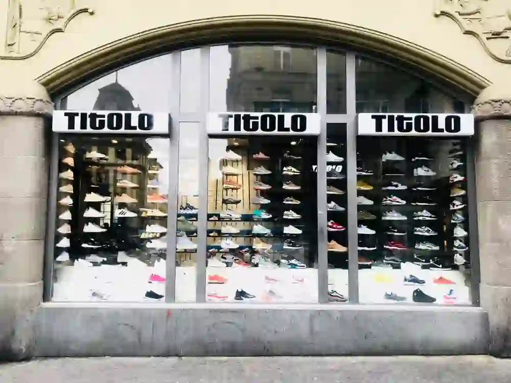Auf dem Bild ist ein Schuhgeschäft zu sehen, das unter dem Namen "TITOLO" betrieben wird. Die großflächigen Schaufenster präsentieren eine umfangreiche Auswahl verschiedener Sneaker-Modelle, die auf mehreren Regalebenen ausgestellt sind. Das Geschäft scheint sich in einem Gebäude mit klassischen architektonischen Elementen zu befinden.