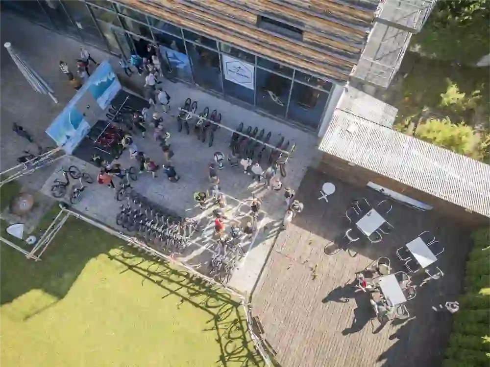 A large group of cyclists ride together along a green forest path. The atmosphere is lively and communal, characterised by movement, closeness to nature and the joy of being on the road together.