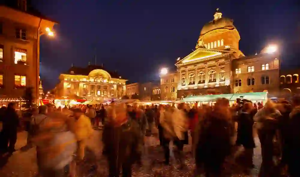 Das Bild zeigt das belebte Treiben des Zibelemärits in Bern bei Nacht, mit Menschenmassen, die im warmen Schein der Lichter vor der prachtvollen Kulisse des Bundeshauses flanieren.