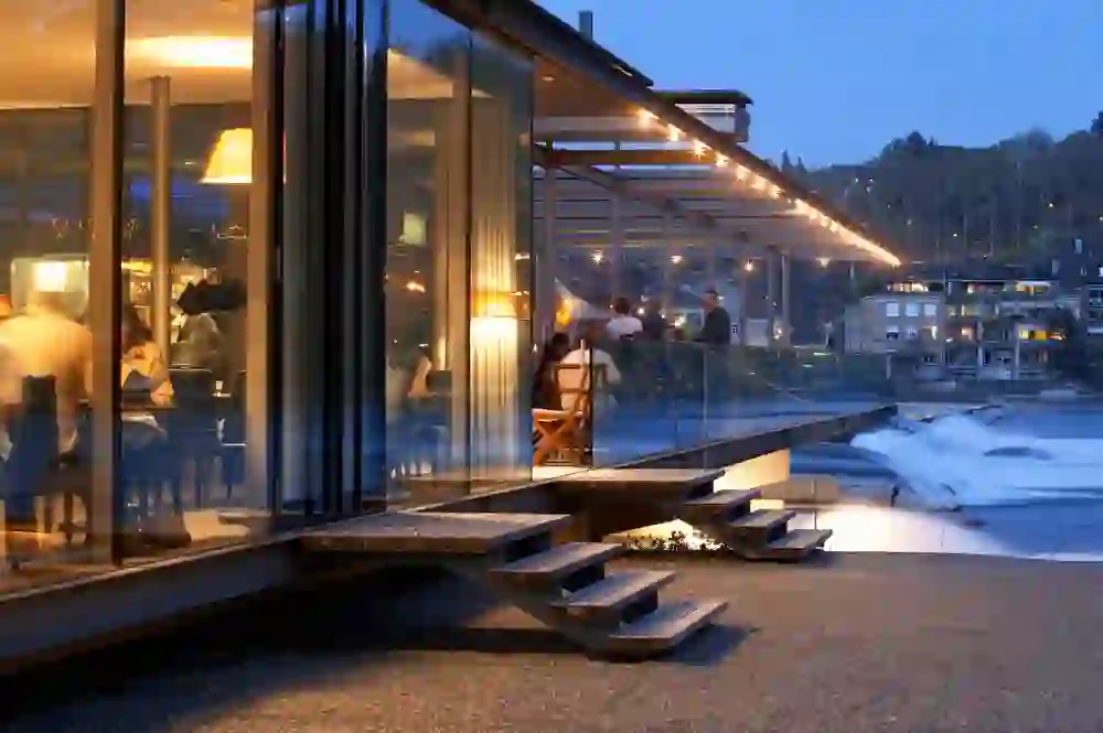 The photo shows the interior of a busy restaurant at dusk with a view of an illuminated outdoor area and a flowing river, the Aare. The warm interior lighting and the string of outdoor lights give the place a cosy and inviting atmosphere. Guests can dine inside or enjoy the view from the terrace. The scene conveys a sense of relaxation and socialising in a picturesque setting.