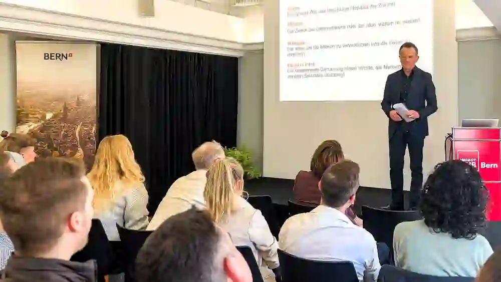 Ein Redner erklärt Konzepte vor einem konzentrierten Publikum. Die Stimmung ist lehrreich und motivierend, während die Zuhörer aufmerksam folgen.