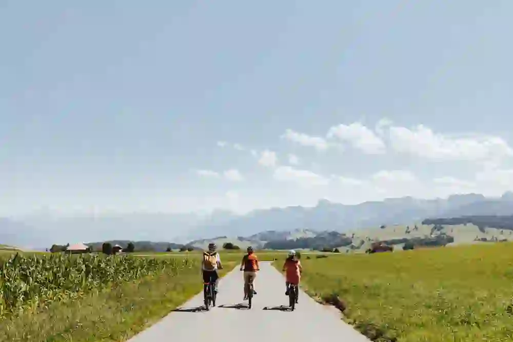 Drei Radfahrer reisen entlang einer ruhigen Landstrasse auf der Sagenroute Gantrisch, umgeben von weiten Feldern, die zum blauen Himmel aufstreben. Im Hintergrund erheben sich majestätische, schneebedeckte Berge. Die Gruppe, angeführt von einer Person im orangefarbenen Shirt, bewegt sich im Einklang mit der friedvollen Landschaft und hinterlässt eine Spur von Harmonie und Entdeckerfreude auf ihrem Weg.