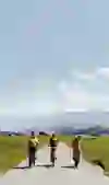 Three cyclists are travelling along a quiet country road on the legendary Gantrisch route, surrounded by wide fields that rise up to the blue sky. Majestic, snow-capped mountains rise up in the background. The group, led by a person in an orange shirt, moves in harmony with the peaceful landscape, leaving a trail of harmony and the joy of discovery along the way.