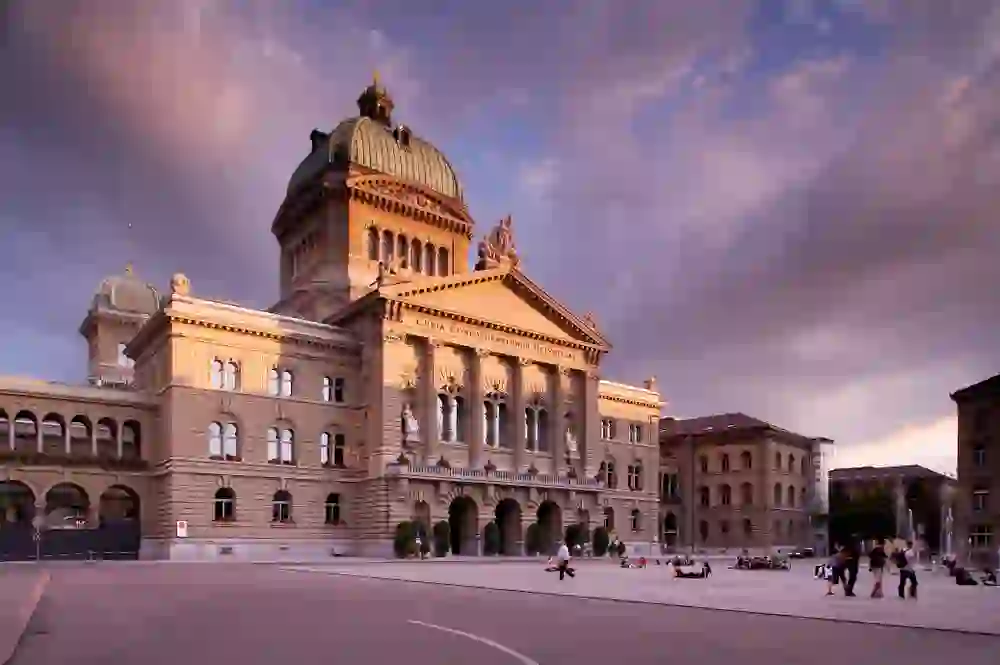 Das Bild zeigt das Bundeshaus in Bern während des Sonnenuntergangs, dessen Fassade von einem warmen, sanften Licht beleuchtet wird, was der Szene eine stimmungsvolle und ruhige Atmosphäre verleiht.