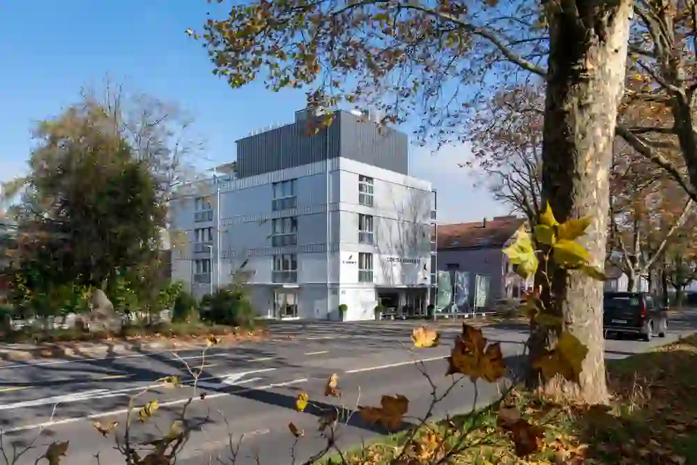 A modern building on a quiet street, framed by autumnal trees and falling leaves. The scene appears clear and urban, yet warm and serene - it conveys stability, calm and a sense of arrival.