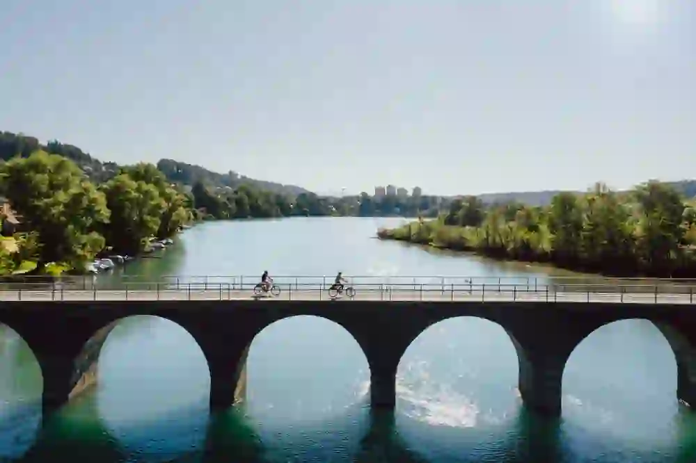 Auf dem Bild ist die Wohleibrücke über den Fluss Aare zu sehen, auf der zwei E-Biker fahren. Das klare blaue Wasser spiegelt die Bögen der Brücke. Im Hintergrund erkennt man üppiges Grün und etwas weiter entfernt Gebäude einer Stadt. Es ist ein sonniger Tag, und die Szene vermittelt ein Gefühl von Ruhe und Freizeit.