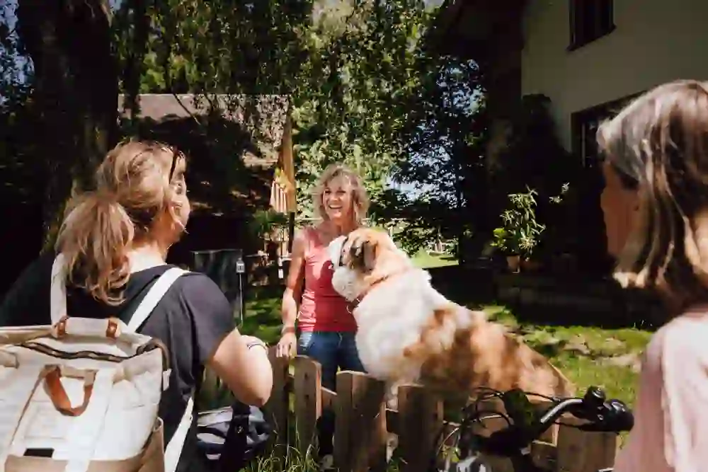 Das Foto zeigt eine heitere Szene mit drei Personen und einem Hund an einem sonnigen Tag auf dem Barry-Hof in Schüpberg. Sie befinden sich im Gespräch an einem Gartenzaun, wobei eine Frau sich freudig zu dem grossen, flauschigen Hund herunterbeugt, der sich zutraulich anlehnt. Die lächelnden Gesichter und die entspannte Körperhaltung signalisieren eine warme und freundliche Begegnung. Ihre E-Bikes im Hintergrund zeigen, dass sie auf der E-Bike Route Fernsichtroute Frienisberg unterwegs sind und eine angenehme Pause einlegen, um den Moment und die Gesellschaft zu geniessen.