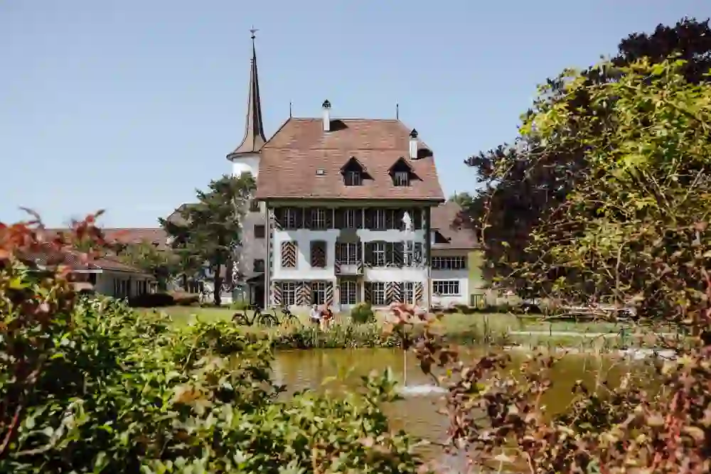 This picture shows Riggisberg Castle in an idyllic garden surrounding the estate. The architecture of the castle, with its characteristic roof shape and striking tower, is classic and inviting.