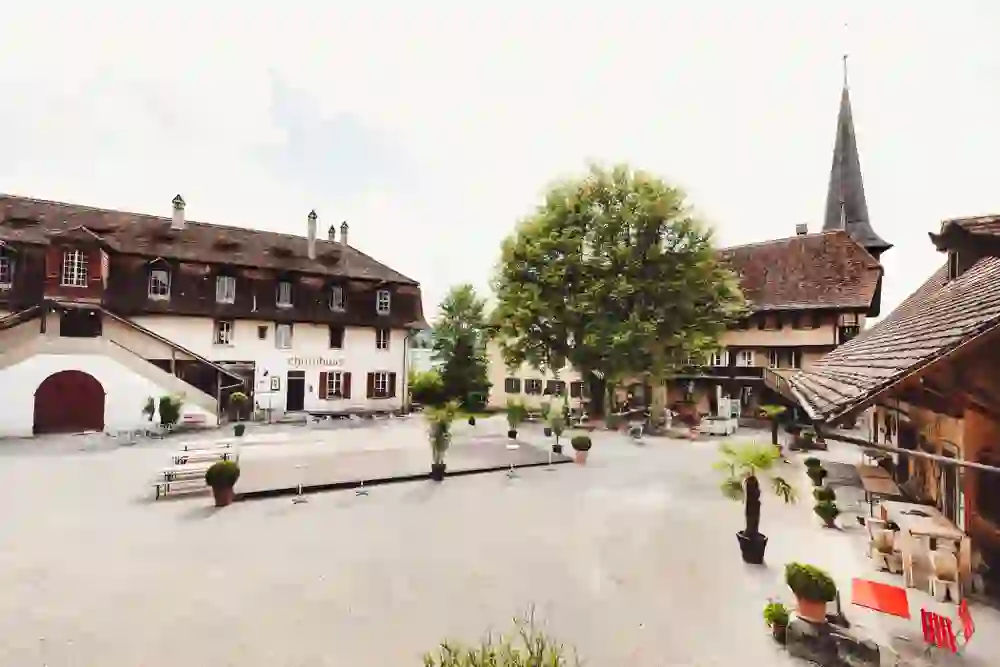 Das Foto zeigt den Kulturhof Schloss Köniz, ein Ort mit historischem Charme und lebendiger Kultur. Man sieht eine Außenansicht mit traditionellen Gebäuden, darunter auch das sogenannte "Chornhuus", und einer weitläufigen Freifläche, die als Terrasse oder Veranstaltungsort dienen könnte.