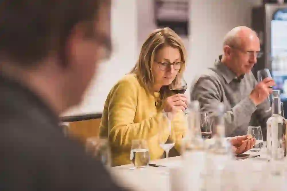 Una donna in giallo si concentra sul profumo del suo vino durante una degustazione, accanto a lei un uomo che degusta. L'atmosfera è rilassata e riflessiva, un momento di piacere e di riflessione.