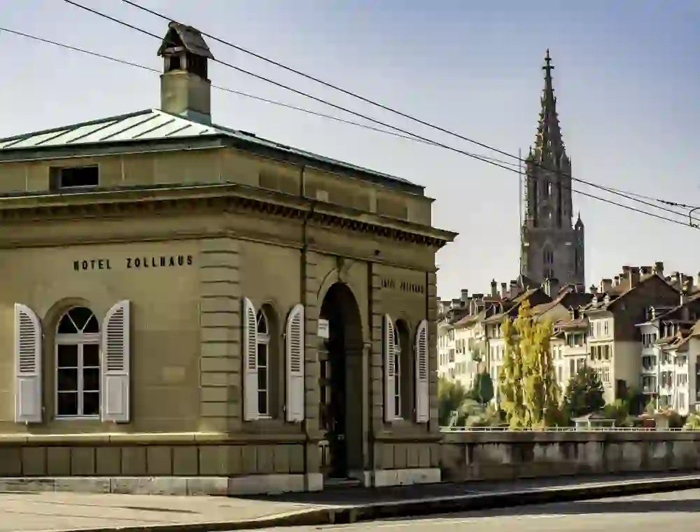 The picture shows the historic customs house in Bern, at the front of the picture, with an impressive view of the Gothic Bern Cathedral in the background, conveying a sense of cultural richness and timeless elegance.