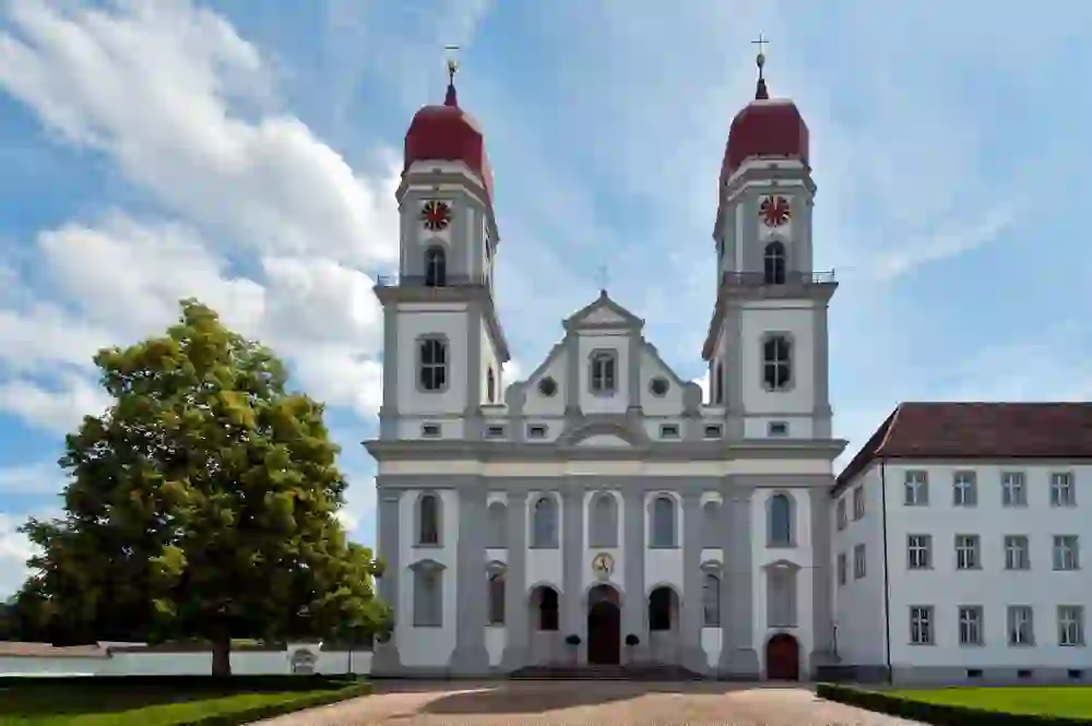 Baroque monastery church with two red towers against a blue sky. The clear façade appears sublime and calm, the spacious grounds radiate dignity and timeless serenity.