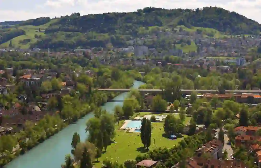 Aus der Vogelperspektive erblickt man das Freibad Marzili in Bern, eingebettet in sattes Grün, mit der Aare, die sich sanft durch die Stadt schlängelt. Die Szene ist eine friedliche Koexistenz von Natur und urbanem Leben unter der Wacht der sanften Hügel im Hintergrund.
