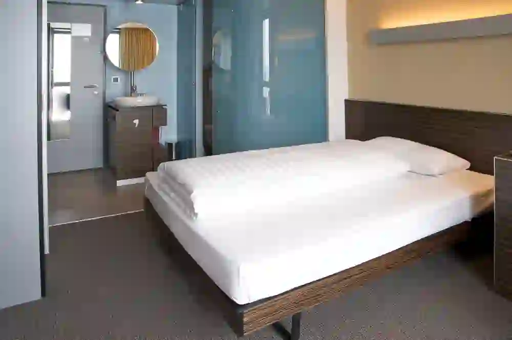The picture shows a modern, minimalist hotel room in the Hotel Ambassador, which radiates a calm and inviting atmosphere. The soft light and clean lines promote a sense of order and relaxation.