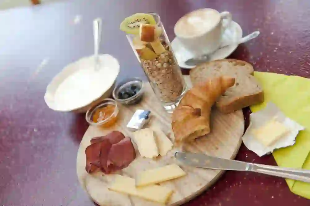 L'immagine mostra una tavola da colazione riccamente imbandita: un barattolo di muesli e frutta fresca, un piatto di yogurt, un croissant, fette di pane integrale, formaggio, salsiccia, marmellata, burro e una tazza di cappuccino. Tutto è disposto in modo appetitoso su una tavola di legno.